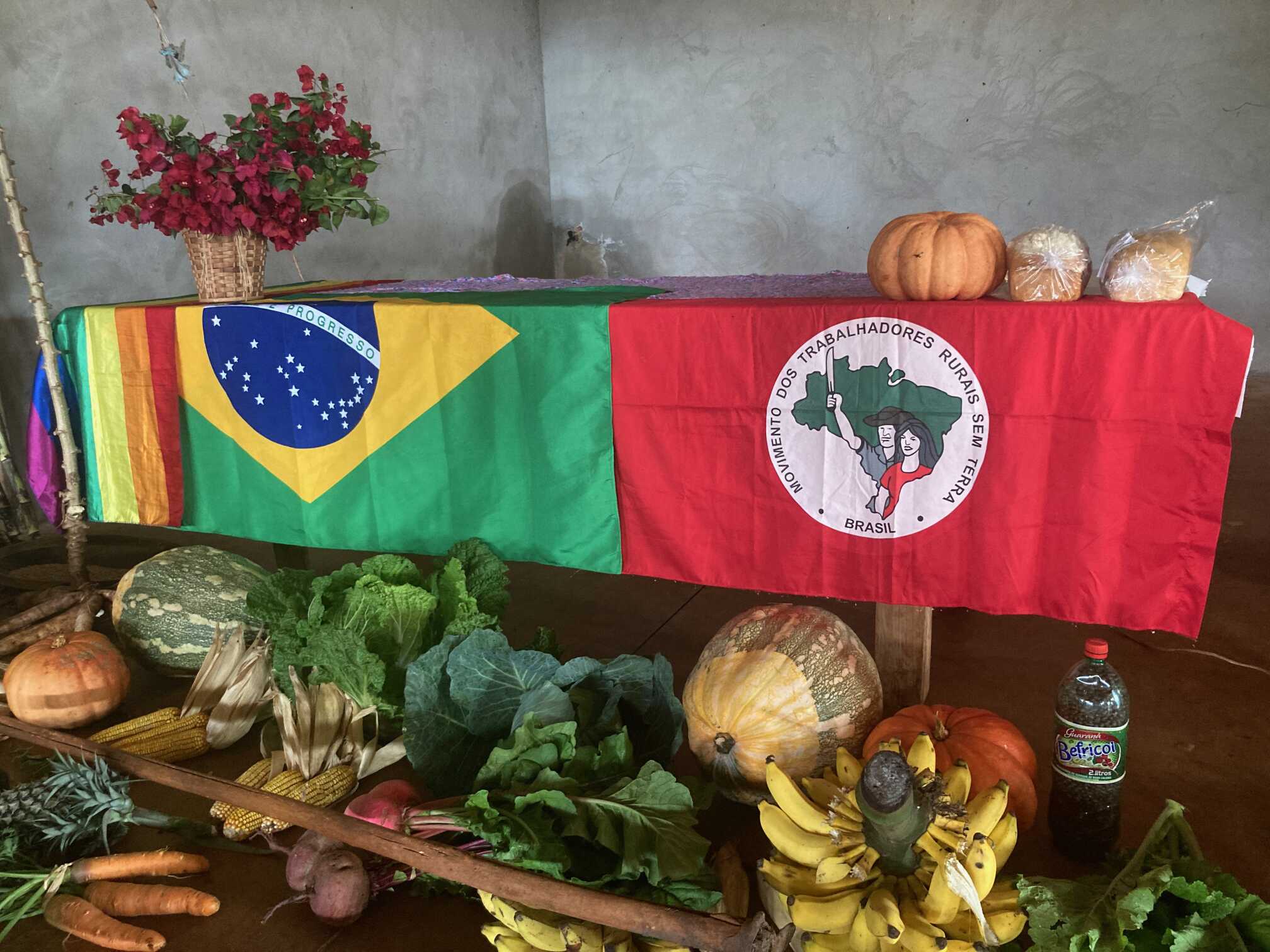 Uma mesa com a bandeira LGBT+, a bandeira do Brasil e a bandeira do MST com várias comidas embaixo, como abóboras, banana, couve, milho etc